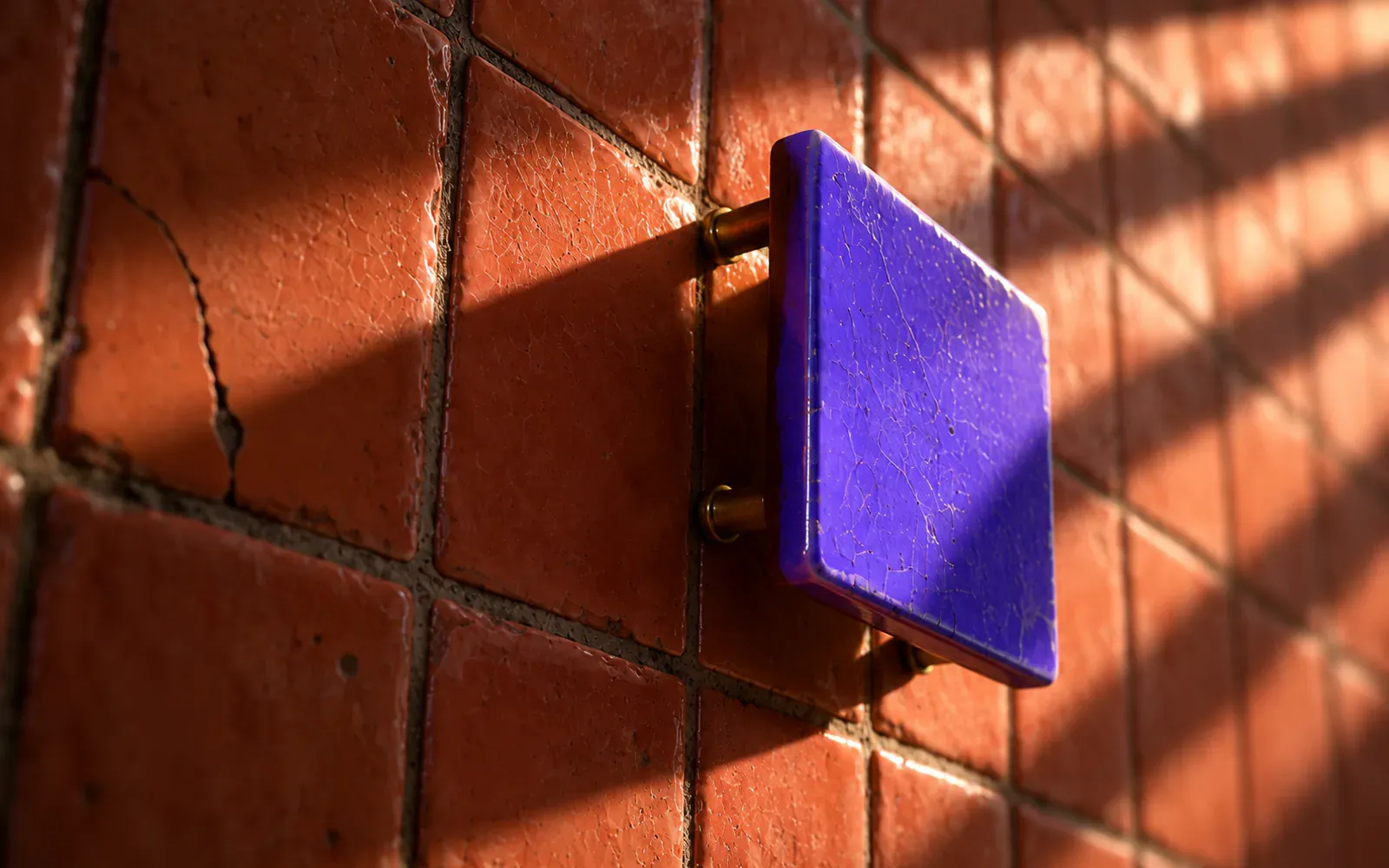 A glossy violet plaque mounted clear of a terracotta tiled wall, hard shutter light raking across the tiles behind it.