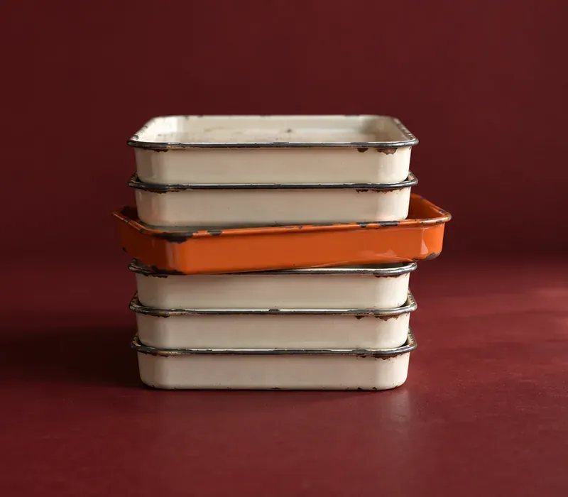 A stack of six enamelled steel trays on an oxblood ground, five bone-white with chipped rims, the third from the top drawn out of the column and glazed orange.